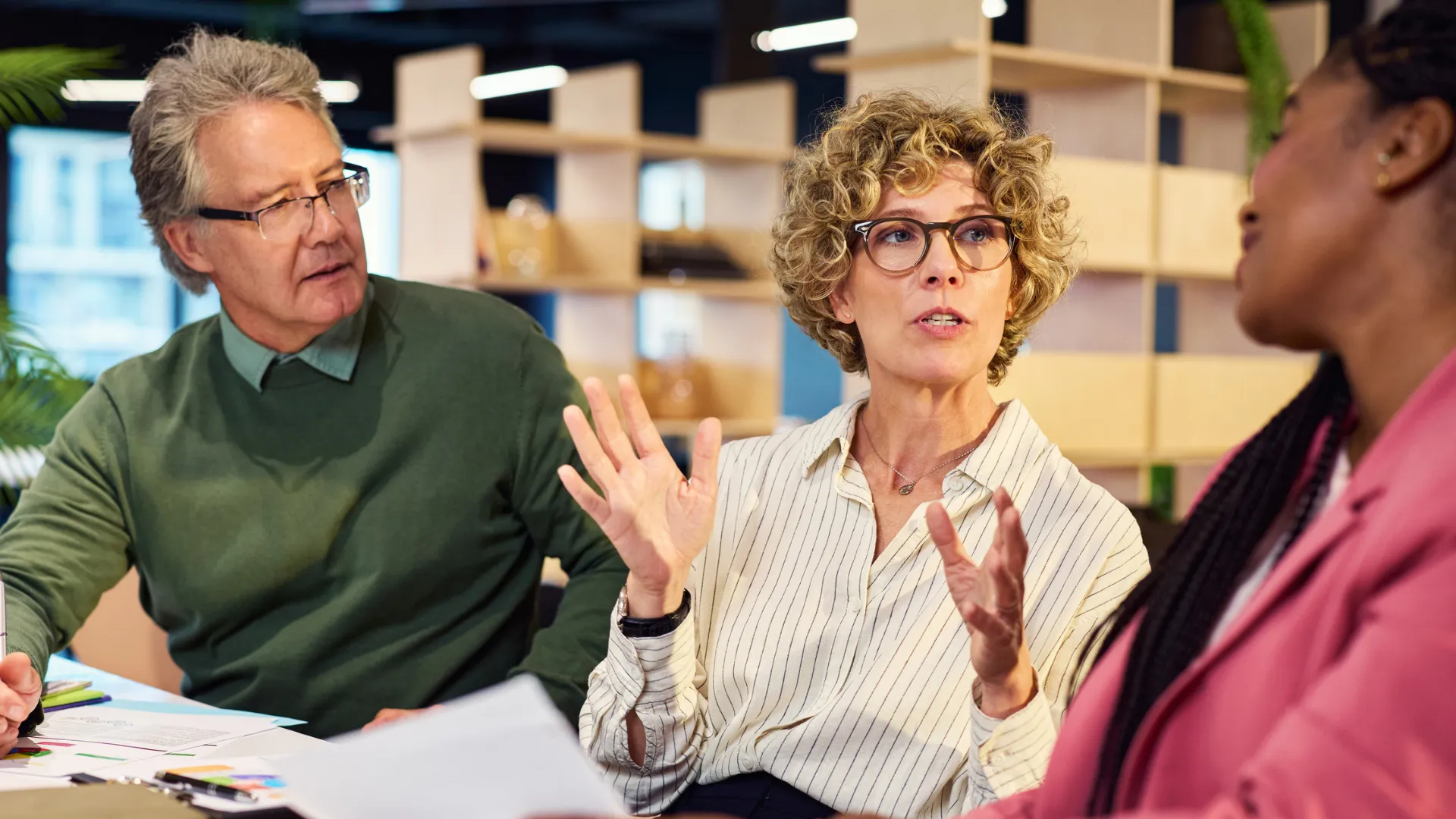 Three professionals in a modern office in an engaged conversation, one speaking expressively with her hands while the others listen.