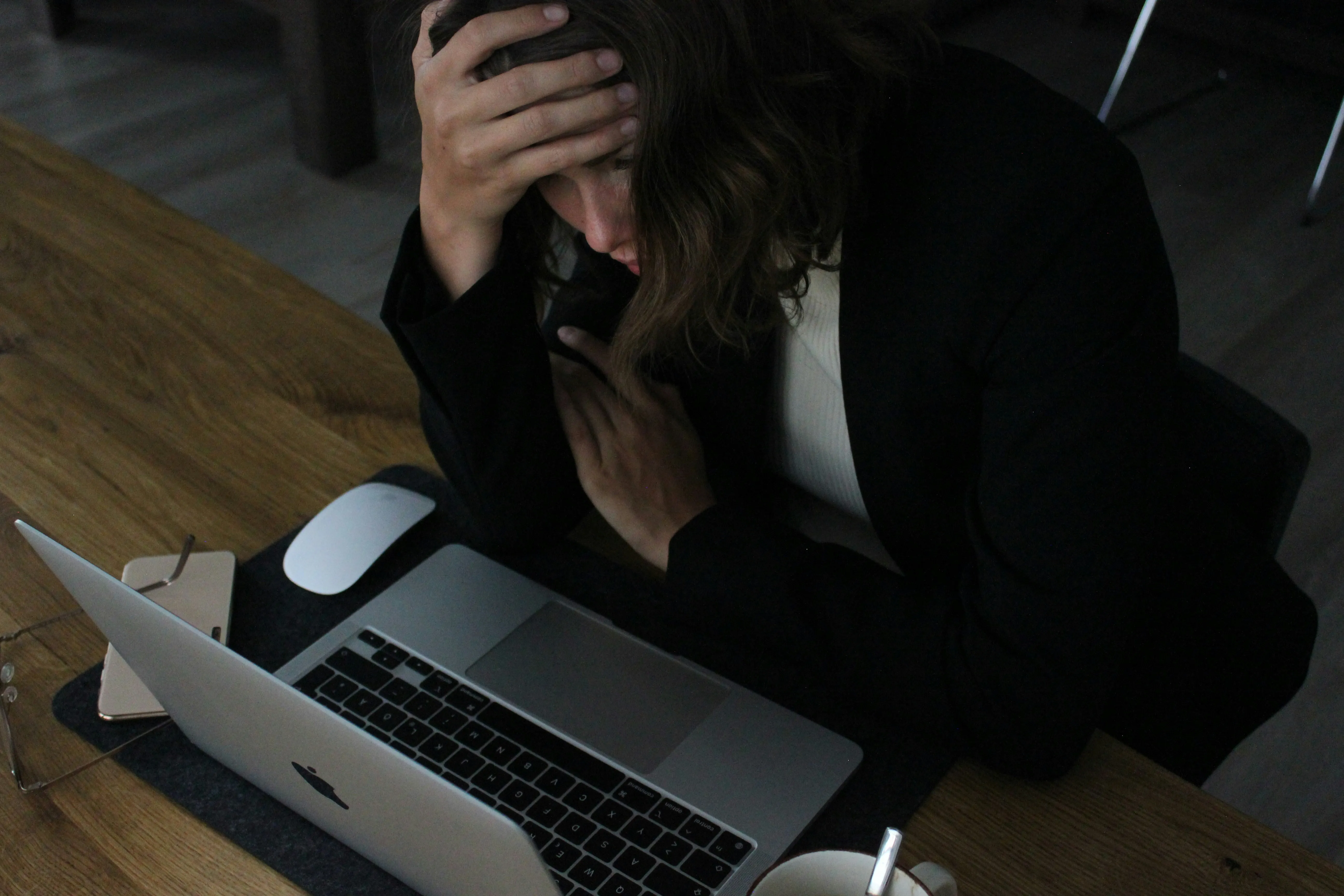 A woman in a dark blazer sits at a desk with a laptop, one hand on her forehead and the other on her chest, looking overwhelmed.