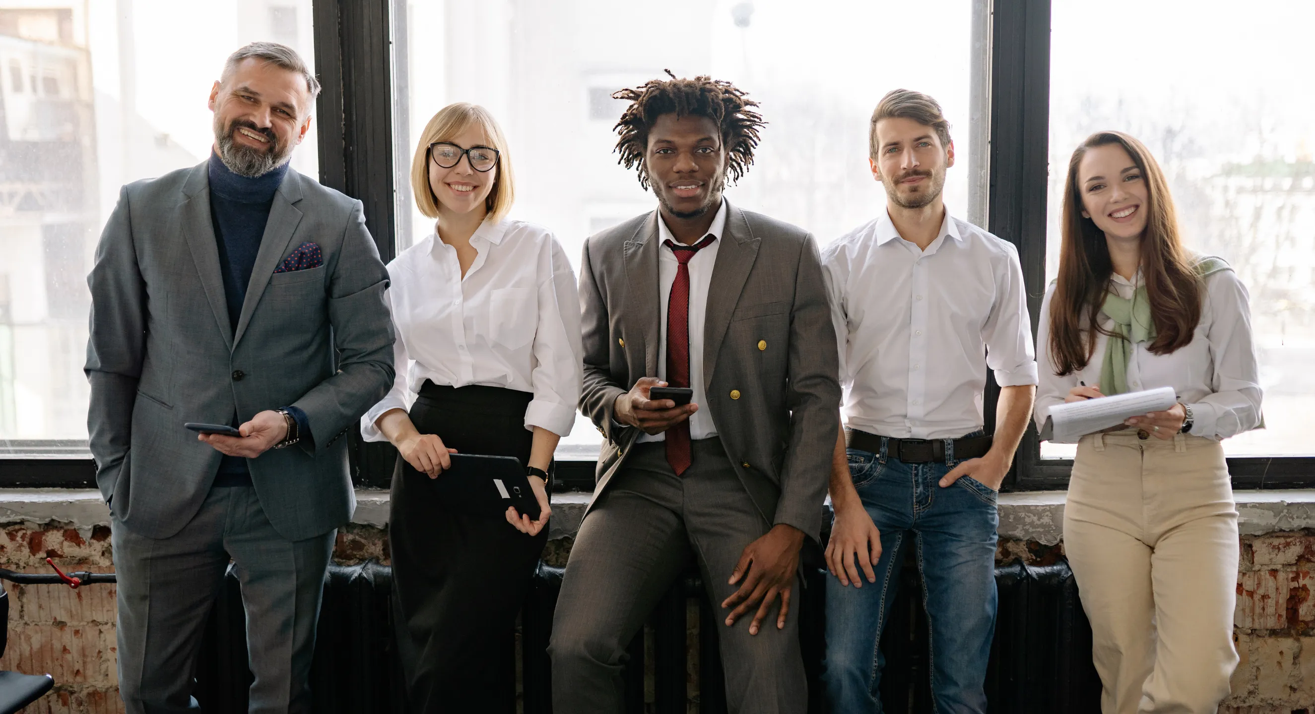 Five diverse professionals stand together and smile by a wall of windows in a bright office.