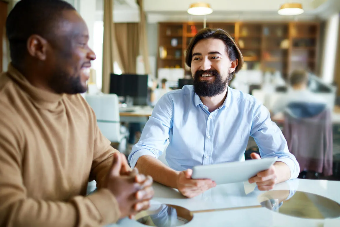 Two colleagues sit in conversation at a white table in a bright open-plan office, one holding a tablet, both smiling.